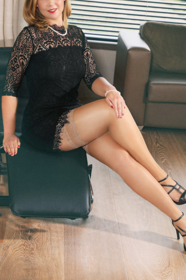 Kira - Escort Lady Duesseldorf im kurzen schwarzen Kleid seitlich auf einem Sessel sitzend.